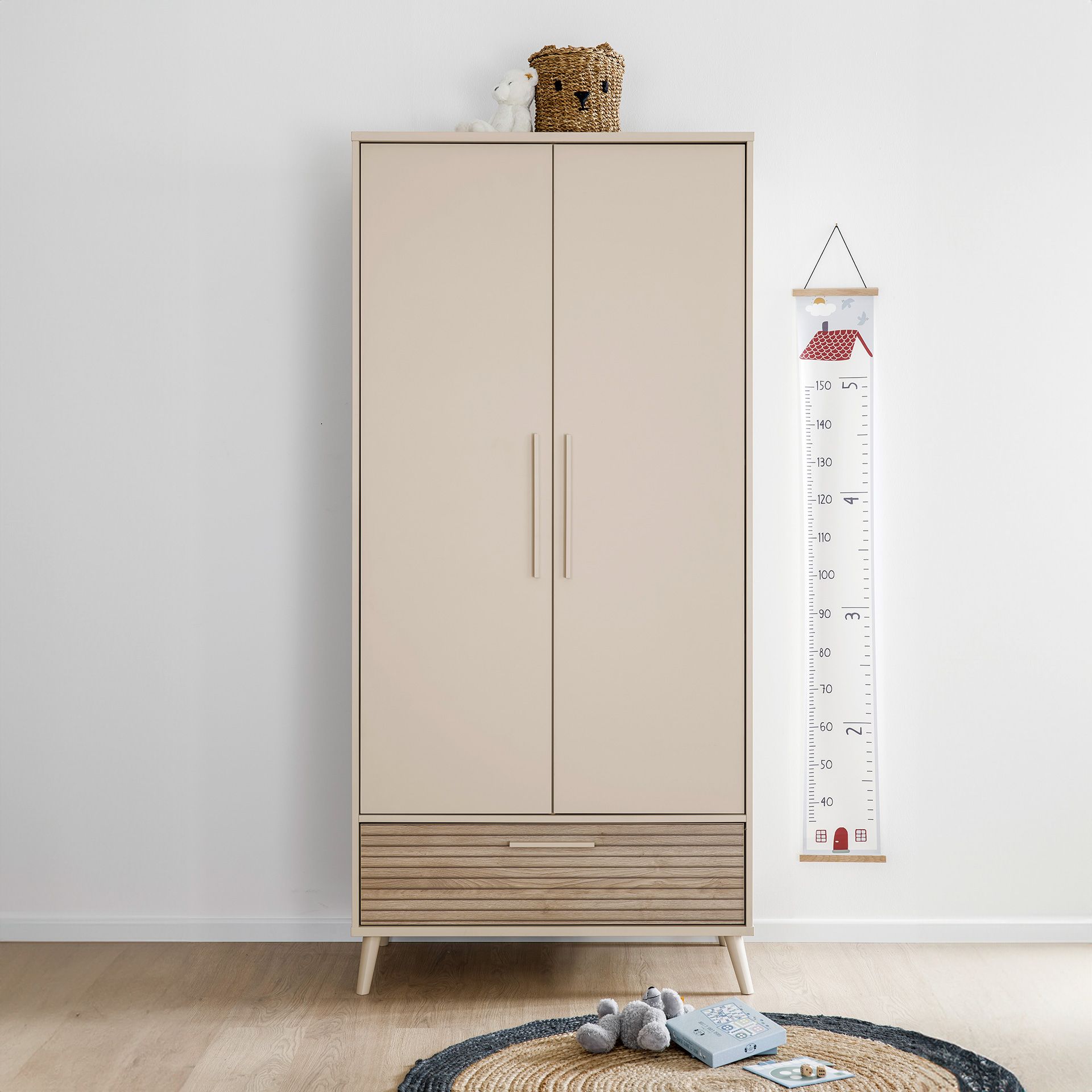 Beiger Kleiderschrank mit Spielzeug darauf, Messlatte an der Wand, Teppich und Spielzeug auf dem Boden.