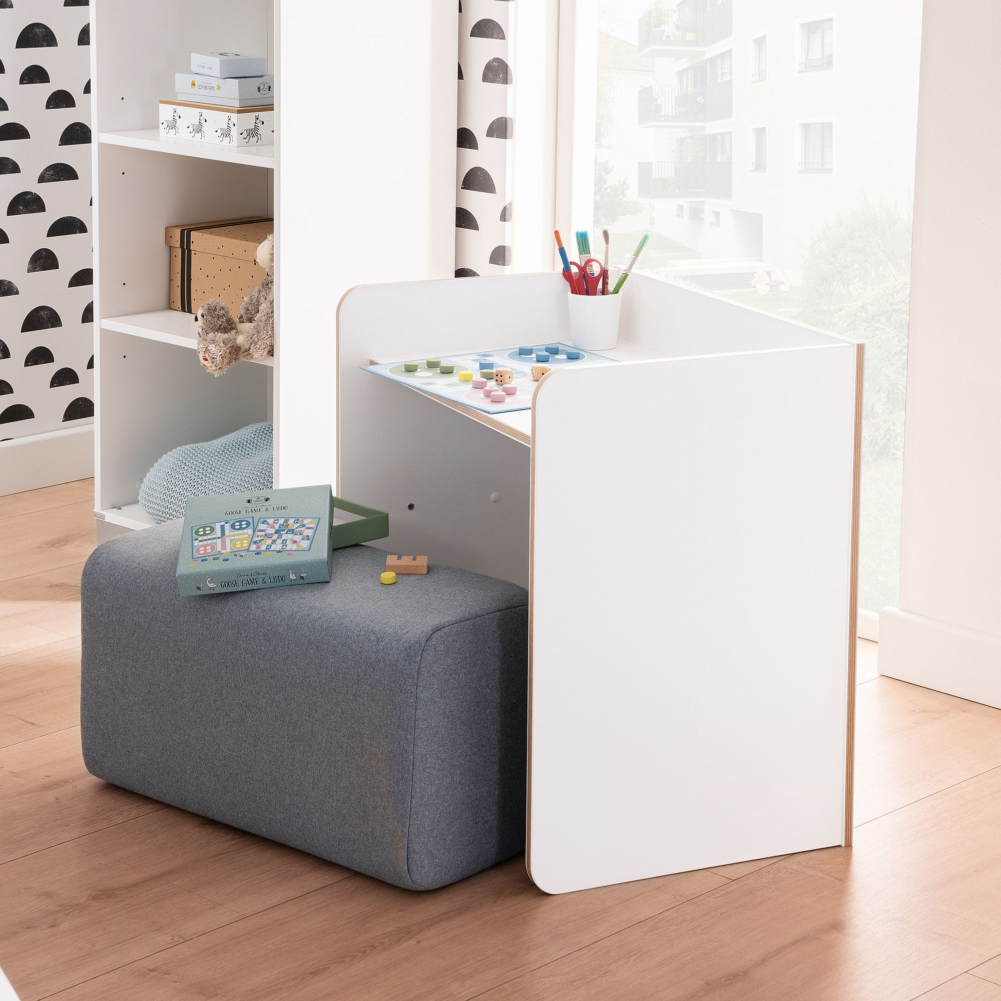 Kleiner weißer Kinderschreibtisch mit Malutensilien und einem grauen gepolsterten Sitz daneben.