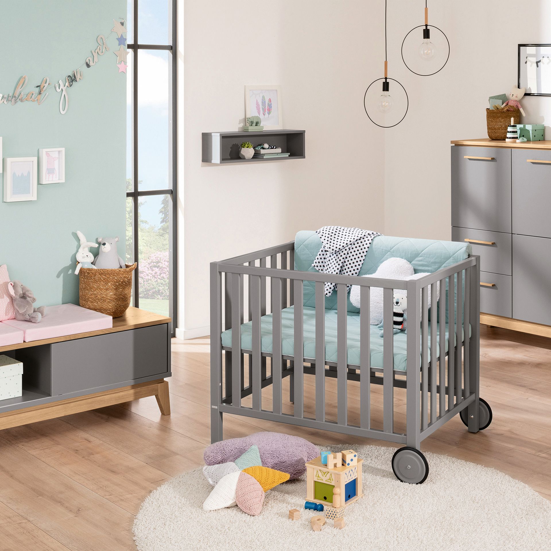 Gray crib in a modern nursery with toys, cushions, and natural light.