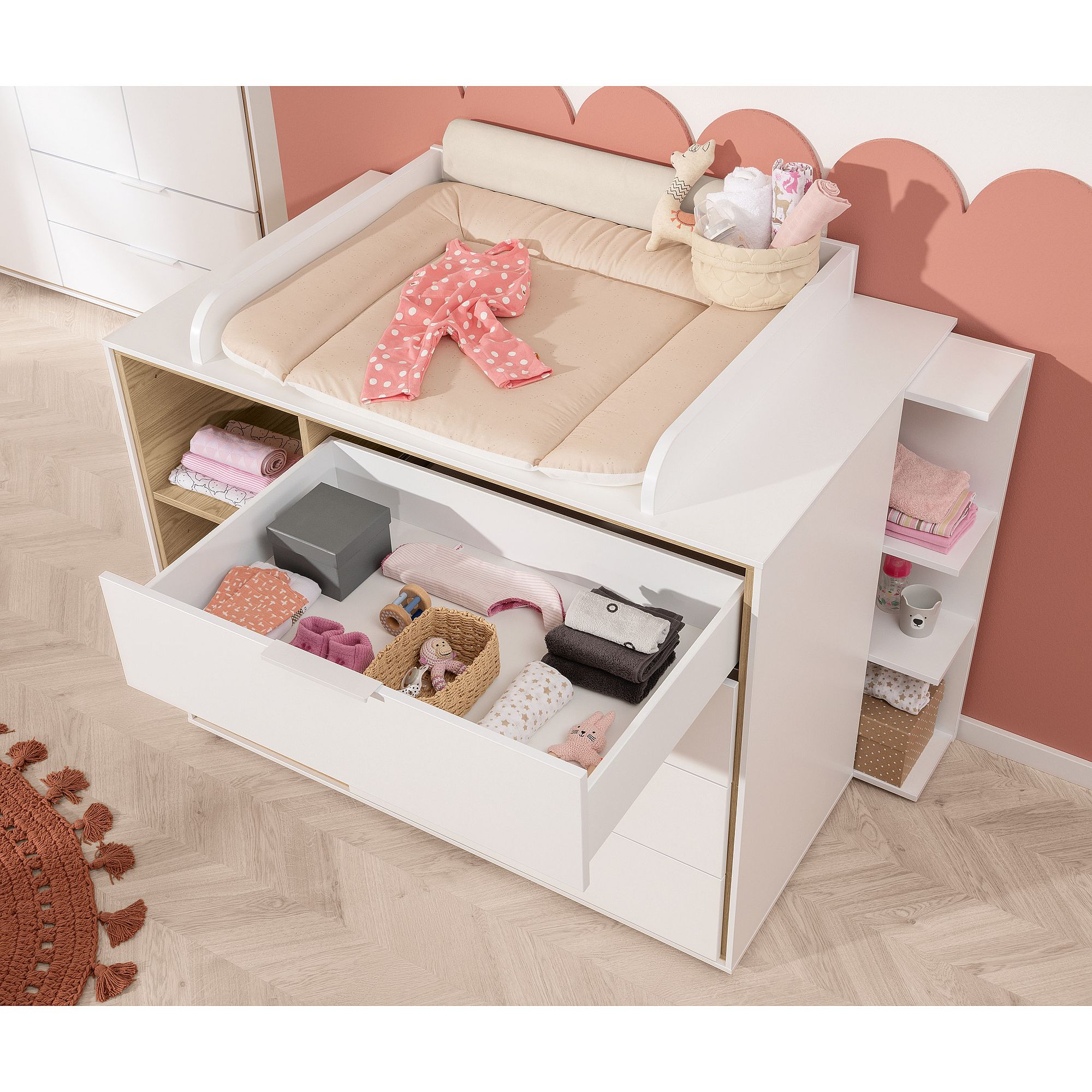 Ein weißer Wickeltisch mit offenen Schubladen, in denen Babykleidung und -zubehör aufbewahrt werden.