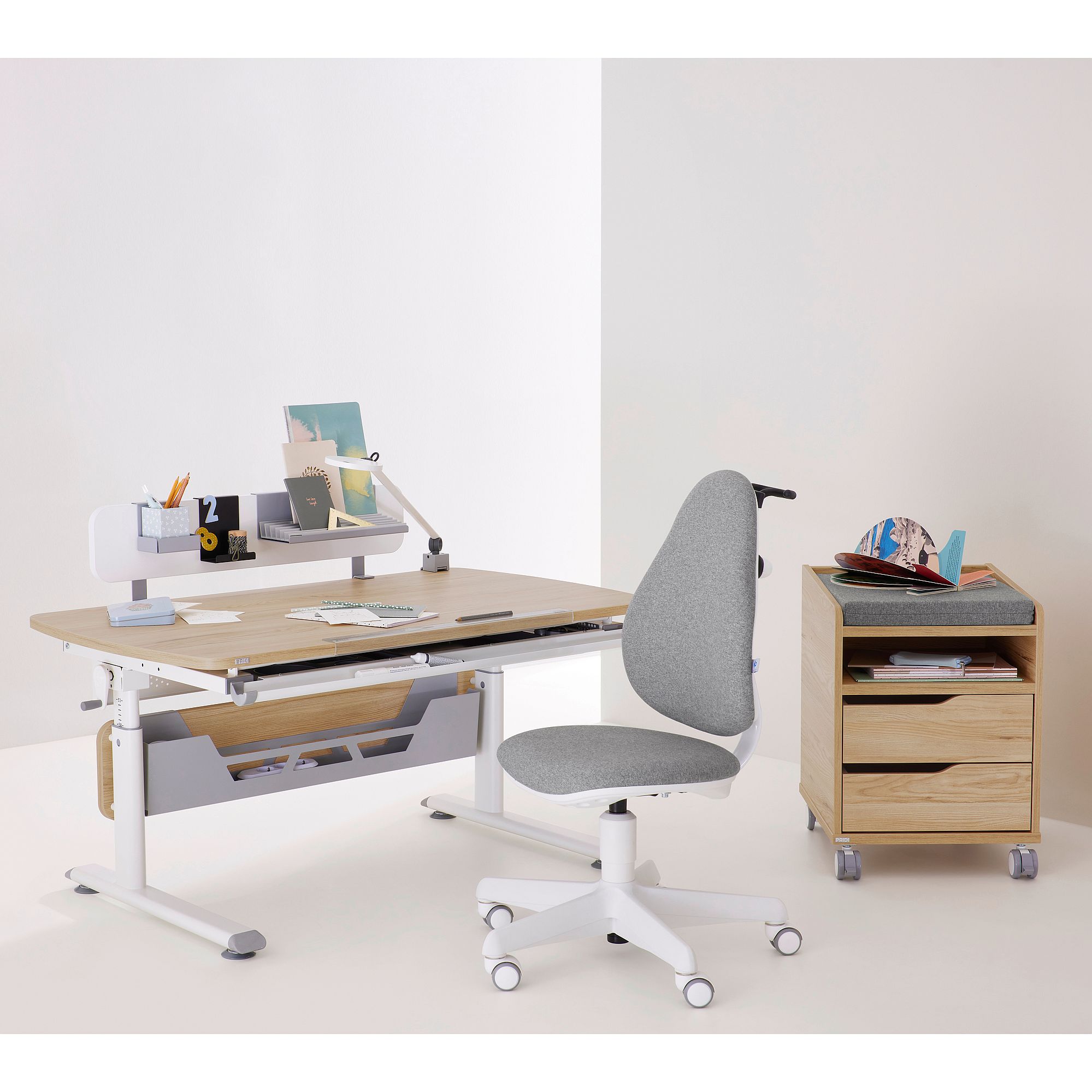 Schülerschreibtisch Diego Curved 120 x 70 cm in Eiche Montana Moderner Arbeitstisch mit Büchern, einem grauen Stuhl und einer Holzschublade auf Rollen.