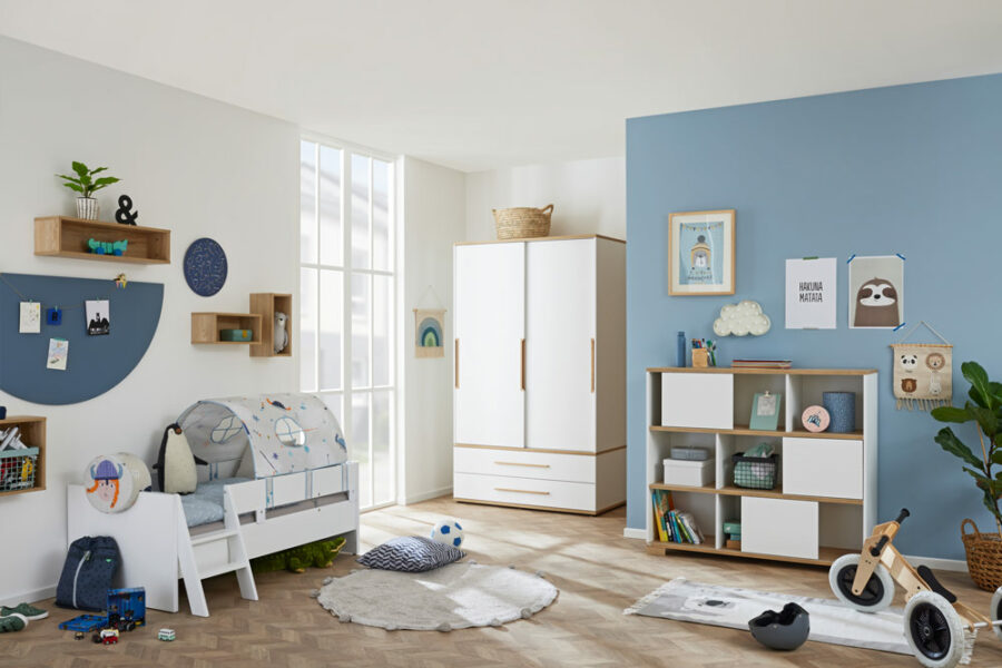 1101_Babyzimmer_paidi_yolanda_junior_0001-900x600-ID1691037_ad4b8841-e391-0690-b42d-4e0ffd097bf3.jpg Helles, modernes Kinderzimmer mit blauem und weißem Dekor, Spielzeug, einem kleinen Bett, Kleiderschrank, Bücherregal und verspielter Wandkunst.