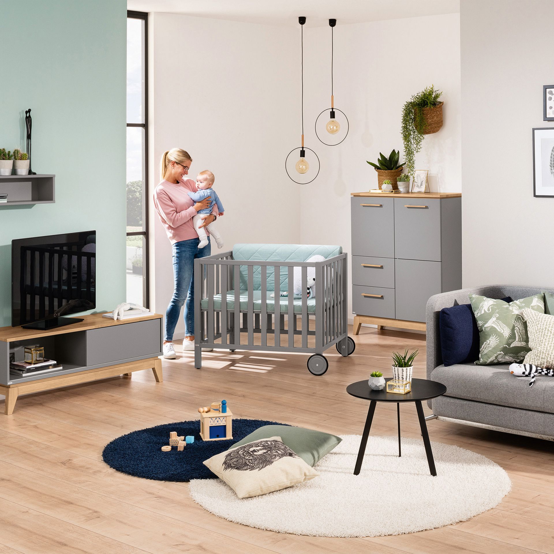 A woman holds a baby in a modern, tidy living room with a crib and toys.