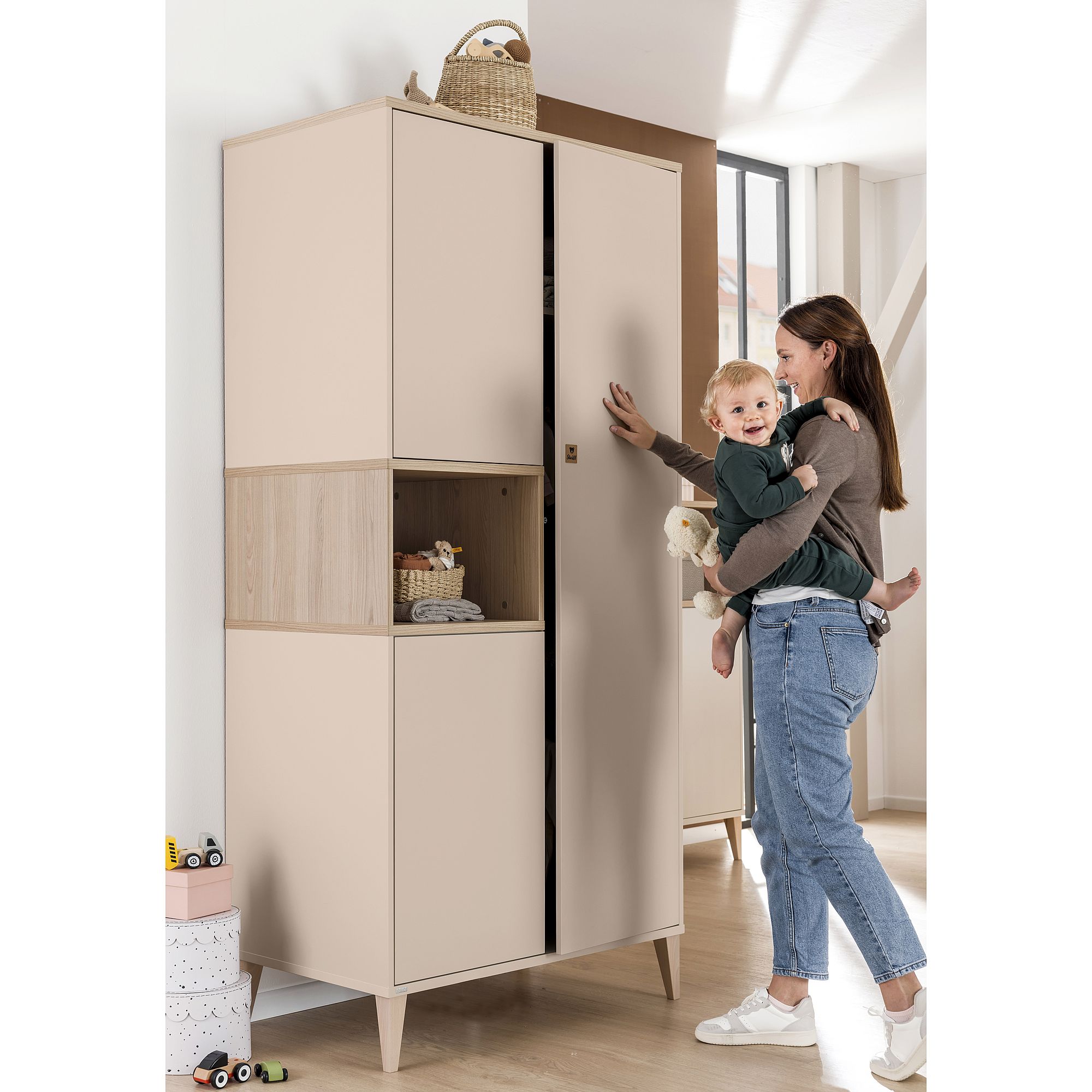 Eine Frau, die ein Kleinkind im Arm hält, öffnet in einem hellen Raum einen beigen Kleiderschrank.