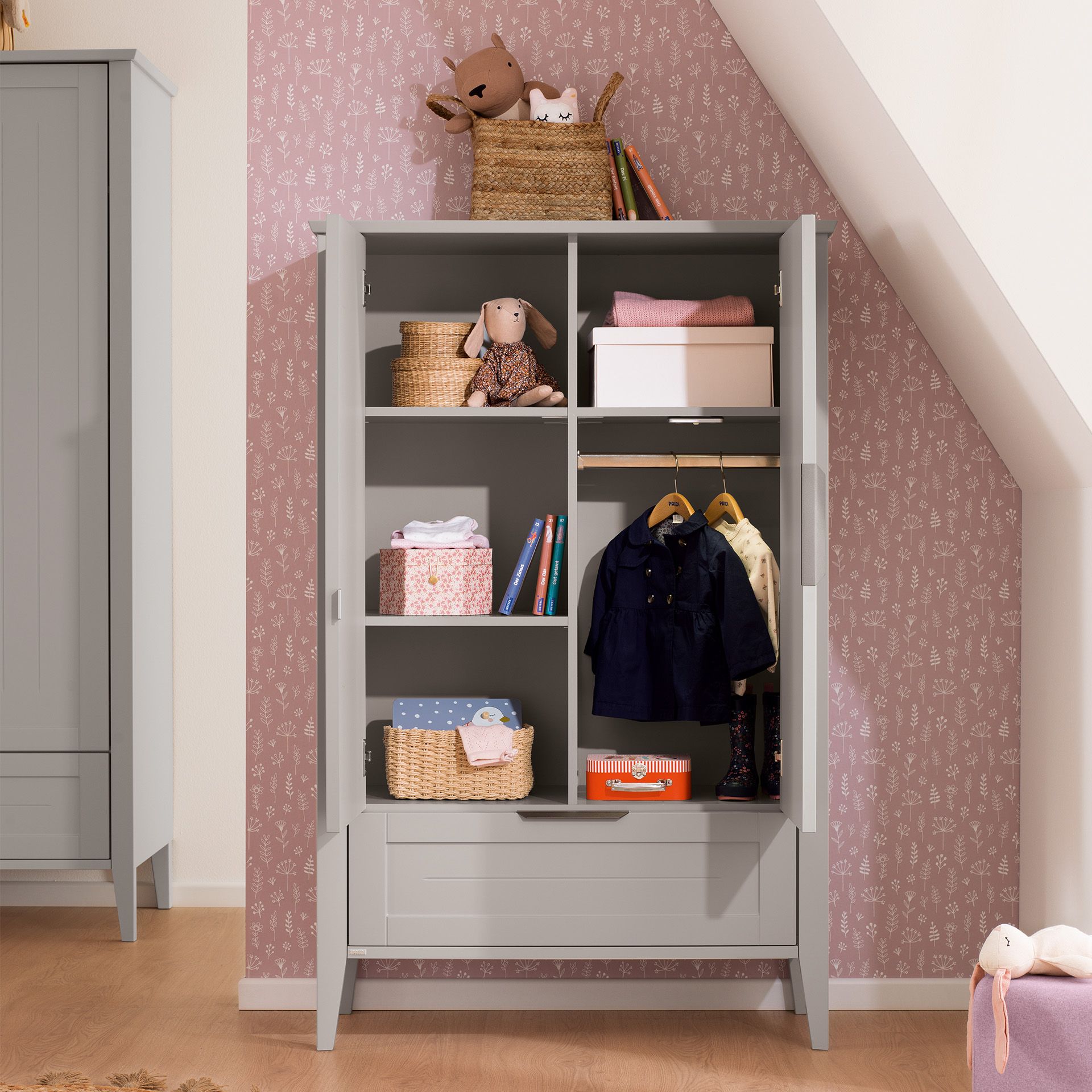 Gray open wardrobe with childrens clothes, toys, and baskets, against pink wallpaper.