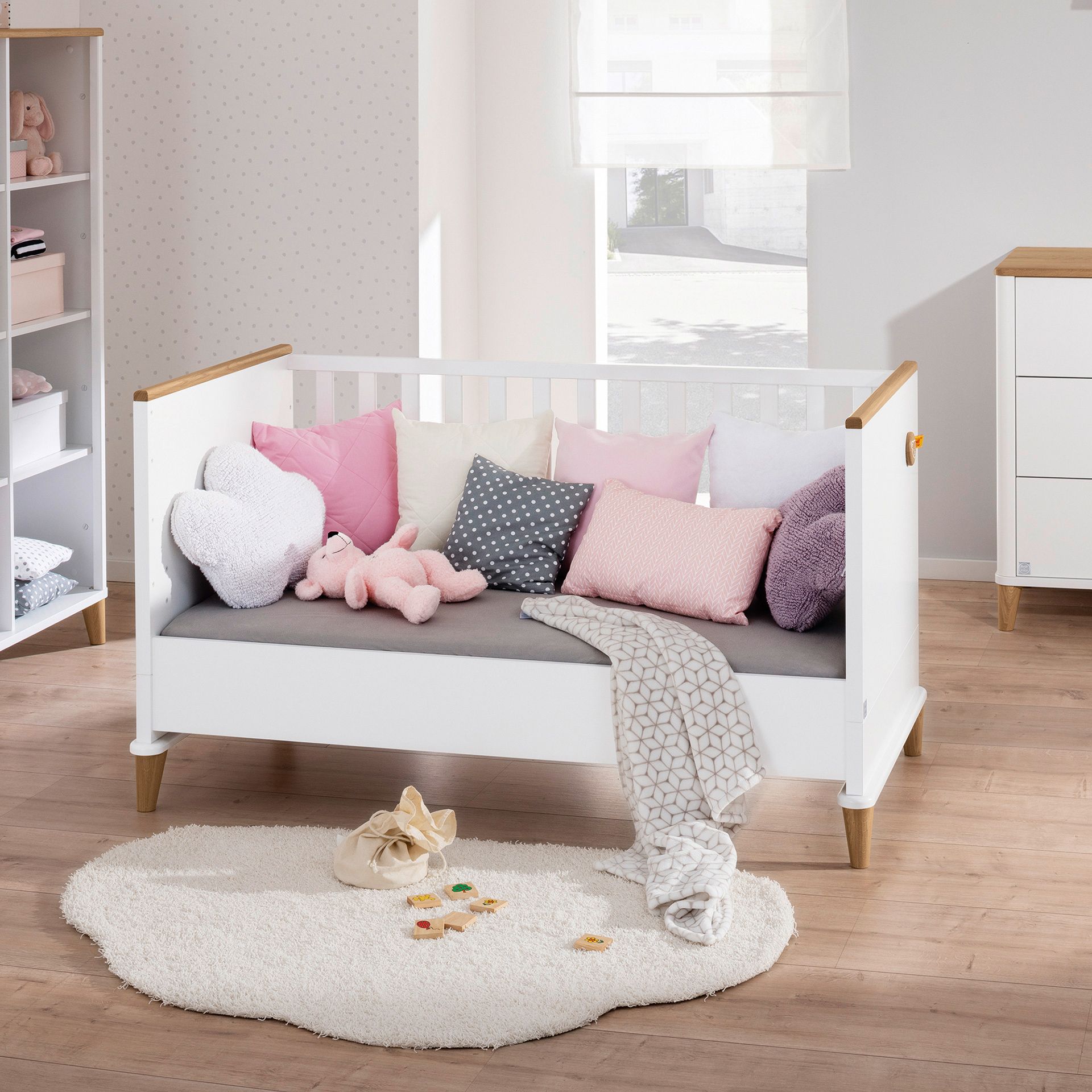 Weißes Kinderbett mit pastellfarbenen Kissen und Spielzeug, in einem hellen, gemütlichen Kinderzimmer.
