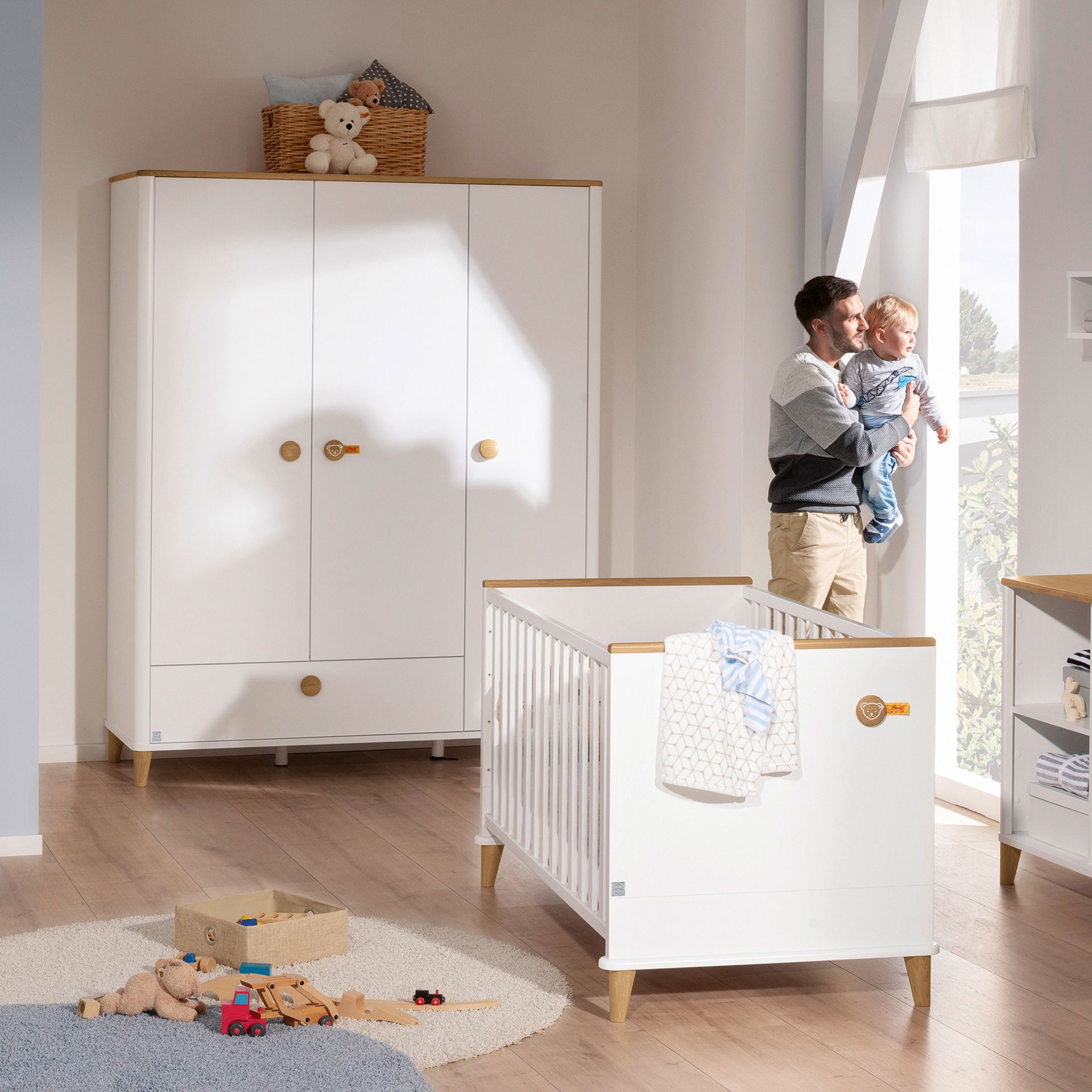 Ein Vater hält ein Baby in einem hellen, modernen Kinderzimmer mit einem Kinderbett und Spielzeug.